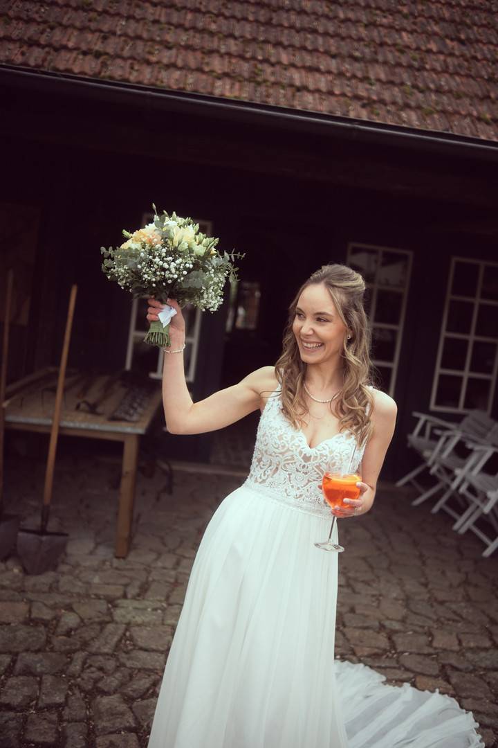 Brautpaarshooting im Antikwerk Breitenthal: Braut mit Brautstrauß und Spritz-Glas lacht auf dem Kopfsteinpflaster